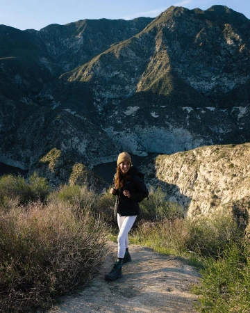 Woman smiles while hiking on a dirt path with mountains in the background on a sunny day with clear skies and green foliage.