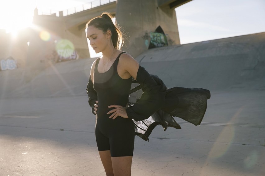 Woman in black bodysuit poses in an urban setting near concrete structures, illuminated by sunlight and wearing long black sleeves.
