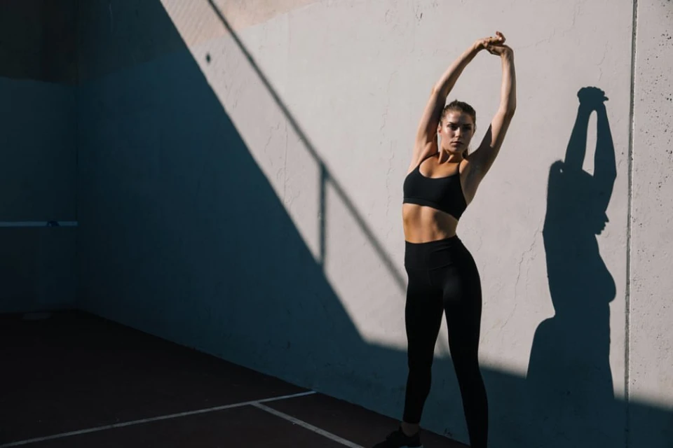 Athletic woman in black workout clothes stretching her arms overhead outdoors in front of a light gray wall with shadows on the surface.