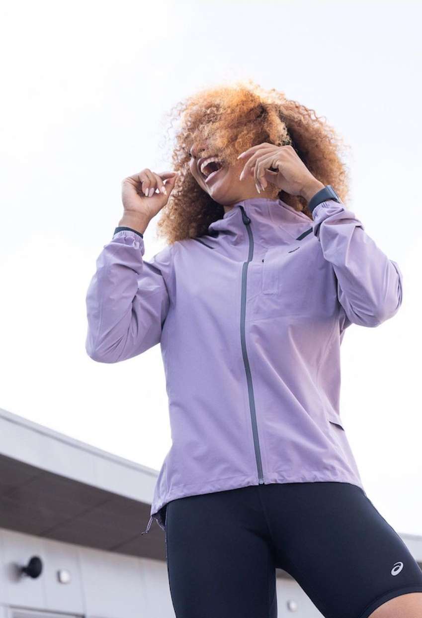 Active woman in purple jacket and black leggings smiles while posing outdoors in natural light, showcasing activewear apparel.