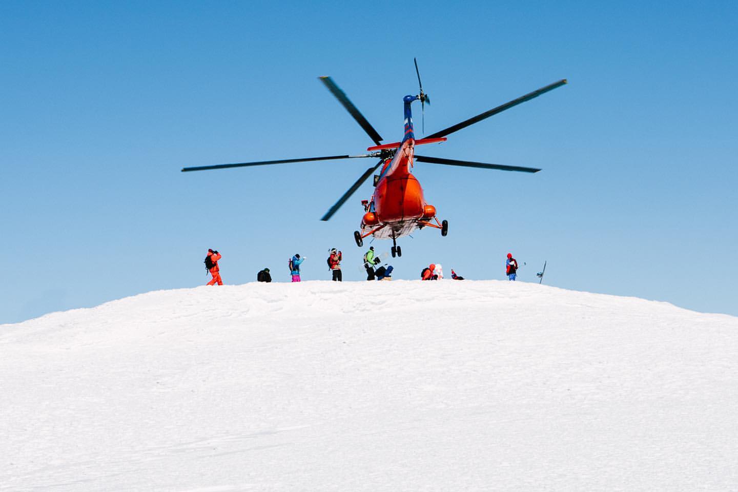 Red and blue helicopter landing on a snow-covered mountain with group of people; clear blue sky above, sunny winter day.