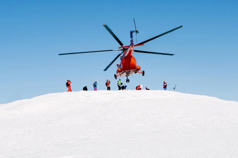 Red and blue helicopter landing on a snow-covered mountain with group of people; clear blue sky above, sunny winter day.