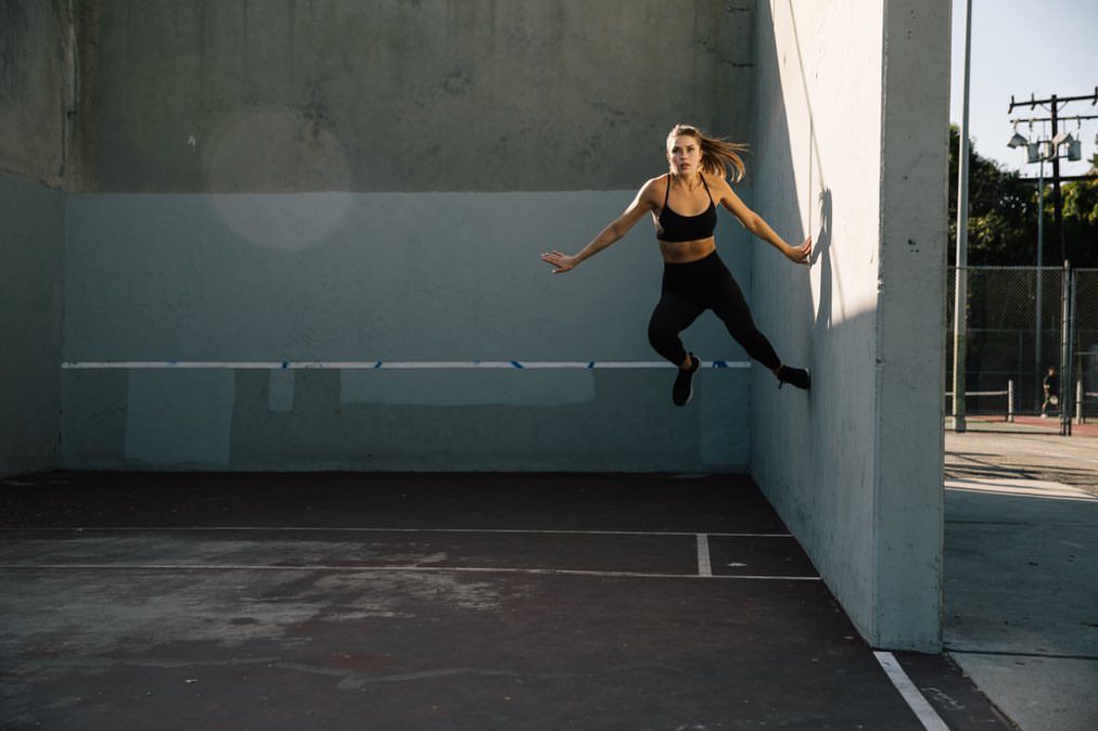 Athletic woman in sports bra and leggings jumps near a concrete wall outdoors, demonstrating fitness and agility in an urban setting.