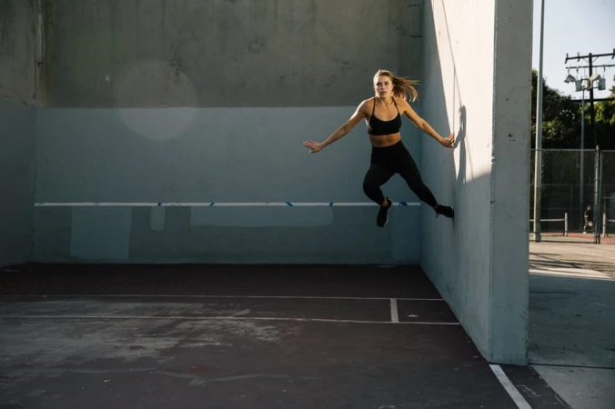 Athletic woman in sports bra and leggings jumps near a concrete wall outdoors, demonstrating fitness and agility in an urban setting.