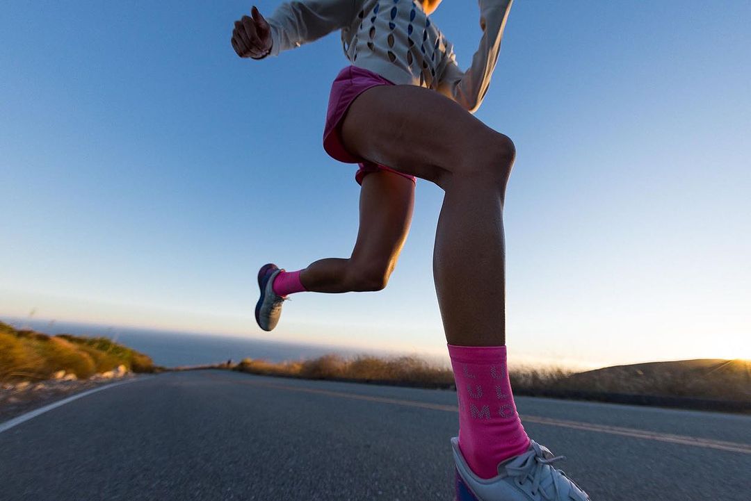 Low-angle view of an adult female jogger running on an asphalt road with ocean and hill views, wearing pink shorts and socks.