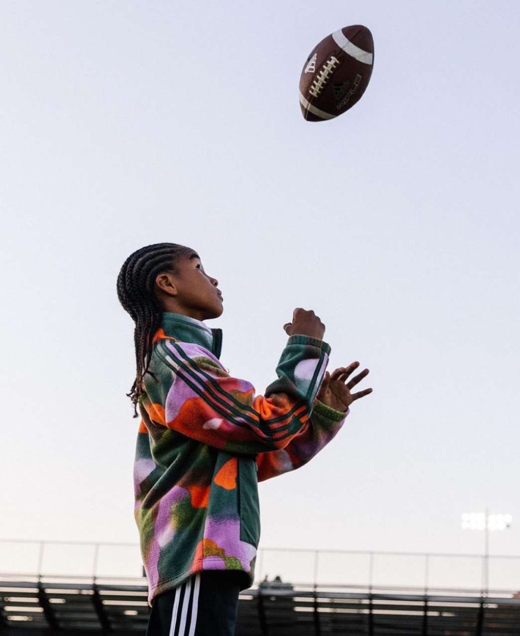 Youthful individual practices football skills outdoors, wearing a vibrant patterned jacket against a clear sky and stadium backdrop.