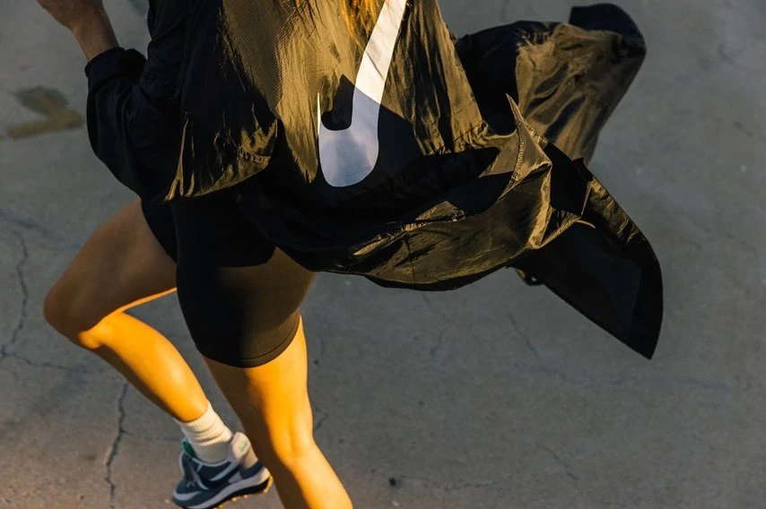 Woman in athletic wear runs on concrete, wearing windbreaker with Nike logo. Close-up, high-angle shot of woman's legs and torso during workout.