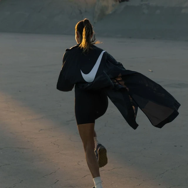 Athletic woman runs outside in sportswear and jacket with Nike logo, captured from behind, on a bright day.