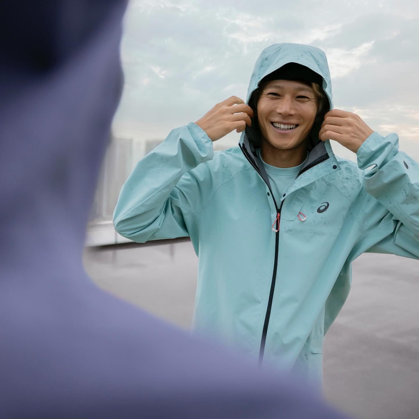 A smiling Asian man in a light blue ASICS hooded jacket adjusting the hood outdoors with a blurred human figure in the foreground.