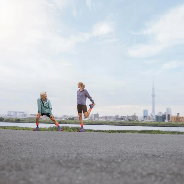 Two women stretch before running on an asphalt path with a distant skyline under a cloudy sky, preparing for a fitness run.