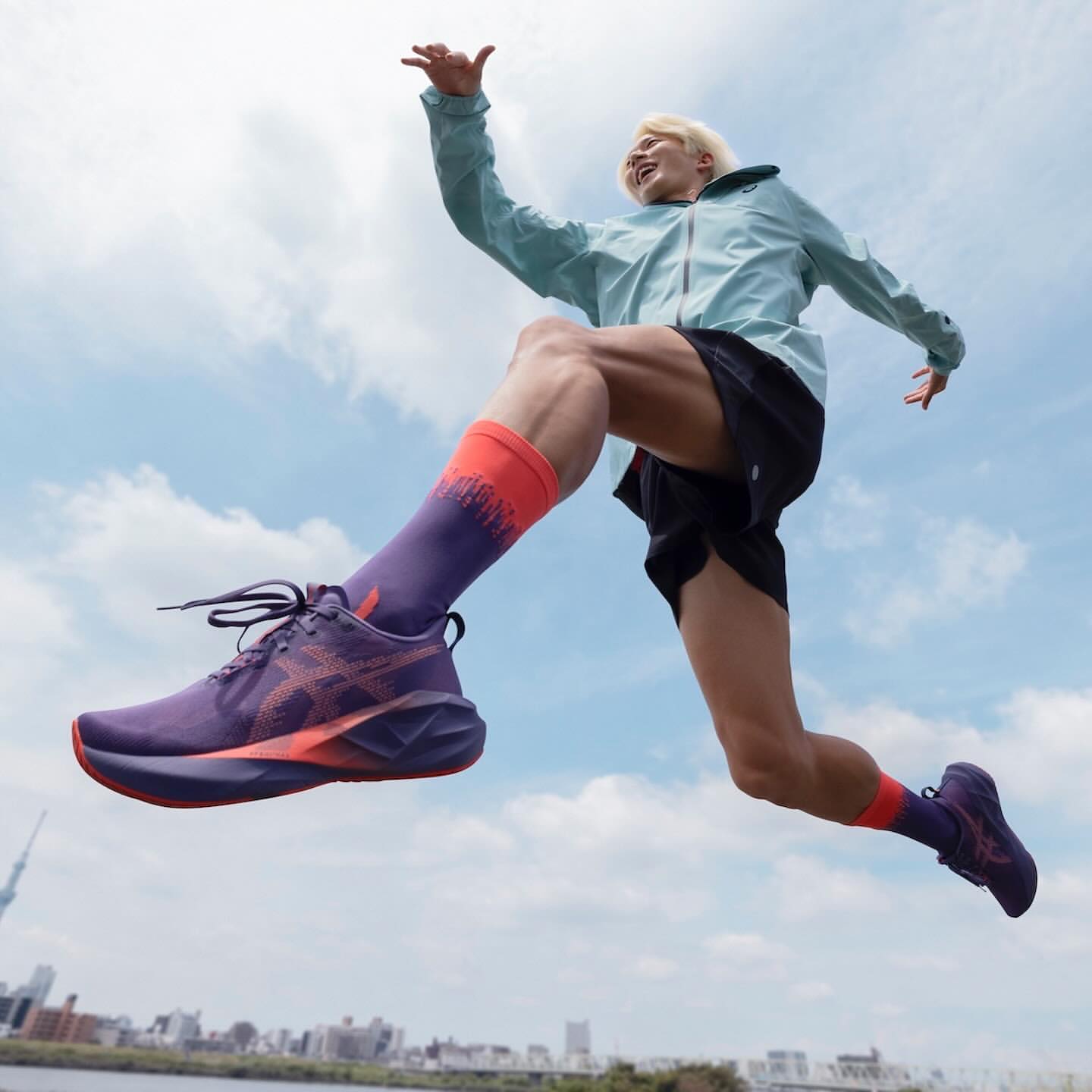 A person mid-jump in athletic wear and running shoes with a purple color scheme with a cityscape and cloudy sky in the background.