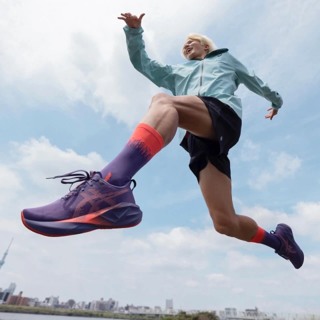 A person mid-jump in athletic wear and running shoes with a purple color scheme with a cityscape and cloudy sky in the background.