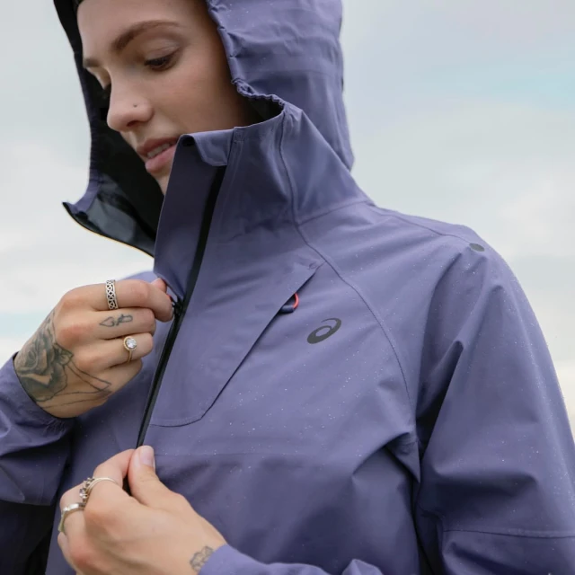 A woman zips up a lavender hooded jacket with the ASICS logo in a close-up shot with a soft cloudy sky visible in the background.