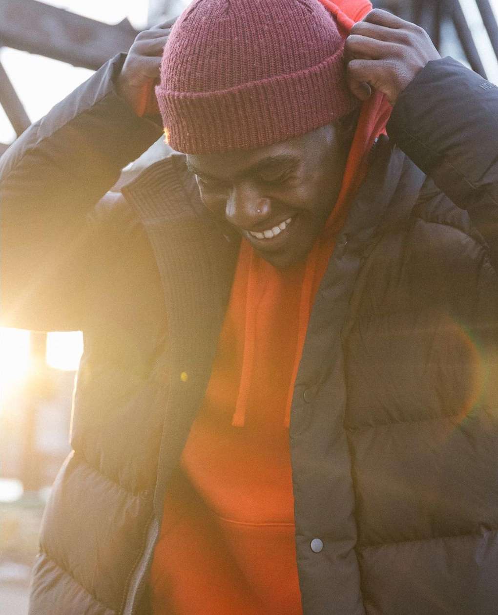 Person adjusts a red beanie and hoodie under a black puffer vest. Outdoor setting with sunlight visible through the background structure.