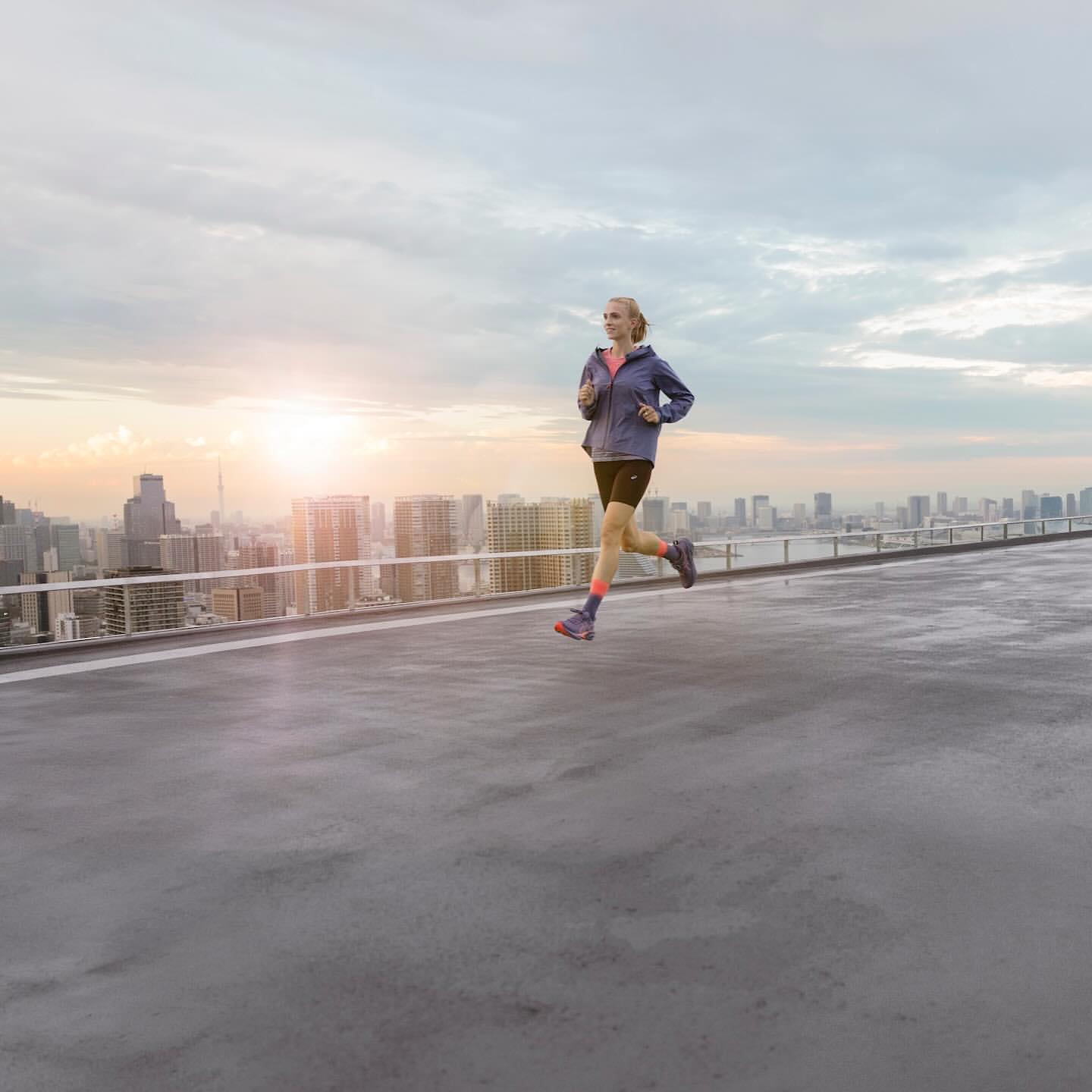 A woman is running on a rooftop in athletic wear. She is jogging on a concrete surface with a cityscape and cloudy sky in the background.