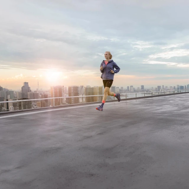 A woman is running on a rooftop in athletic wear. She is jogging on a concrete surface with a cityscape and cloudy sky in the background.