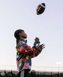 Young Black child with braided hair wearing a patterned fleece jacket, looking up as a football is thrown in the air.