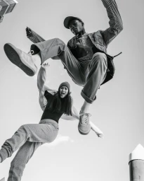 Low-angle black and white action shot of two models jumping mid-air outdoors against a bright sky.