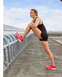 Woman in black athletic top and shorts stretching her leg on a waterfront railing wearing bright red running shoes