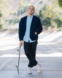 Man standing on a paved path holding a skateboard, wearing a dark cardigan over a light blue button-down shirt.