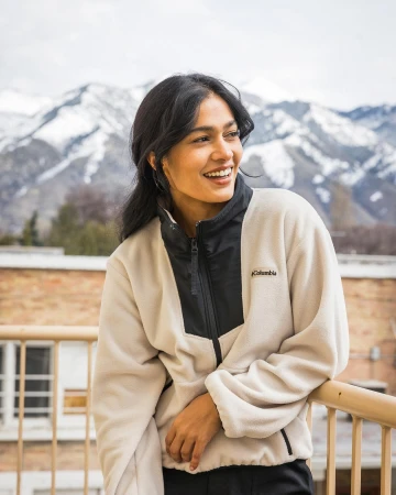 Smiling South Asian woman wearing a cream and black fleece jacket leaning on a railing against a mountain backdrop.