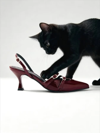 Black cat playfully interacting with a glossy burgundy high-heeled shoe against a clean, bright white backdrop in a studio shot.