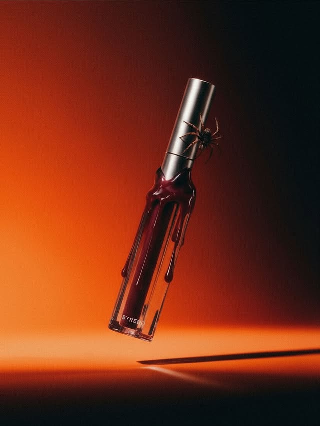 Close-up product shot of a red lip gloss with a clear tube and silver top, featuring a black spider, against a gradient background.
