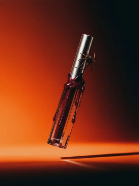 Close-up product shot of a red lip gloss with a clear tube and silver top, featuring a black spider, against a gradient background.
