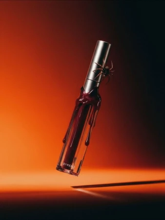 Close-up product shot of a red lip gloss with a clear tube and silver top, featuring a black spider, against a gradient background.