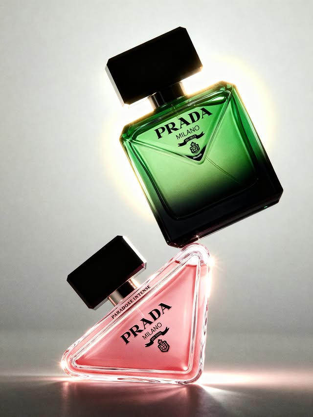 Two Prada Milano perfume bottles, one green and one pink, are stacked at an angle in a beauty advertisement against a grey background.