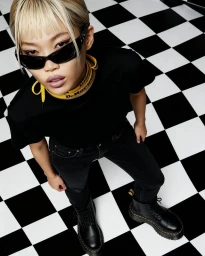 High-angle portrait of an Asian woman with blonde bangs wearing black sunglasses, black t-shirt, dark jeans, and boots on a checkered floor