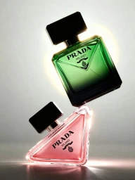 A green glass Prada perfume bottle balanced atop a triangular pink Prada Paradoxe Intense fragrance bottle against a soft grey backdrop.
