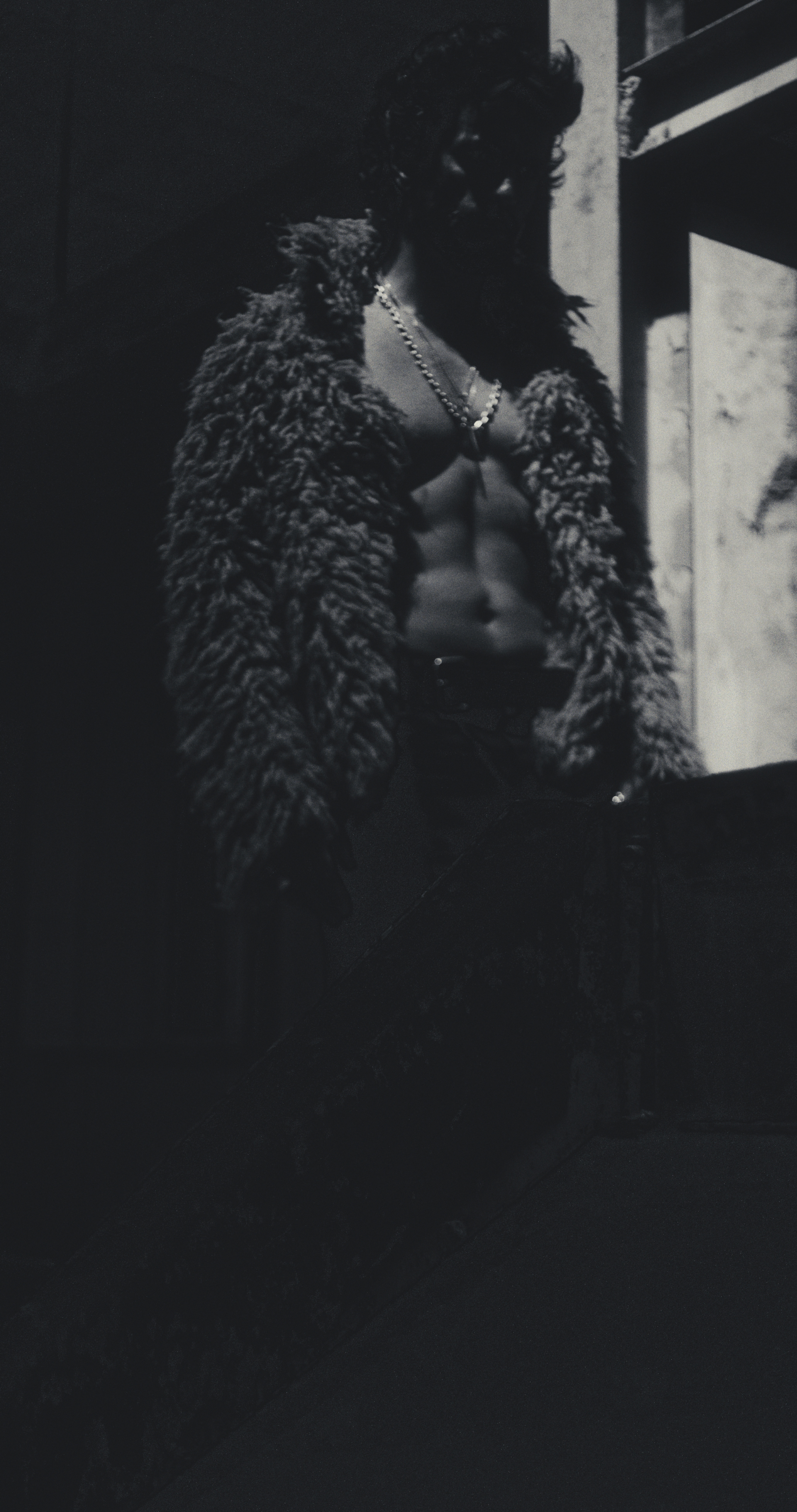 Monochrome portrait of a shirtless man wearing an open shaggy coat and metallic chain necklaces in dramatic shadow.
