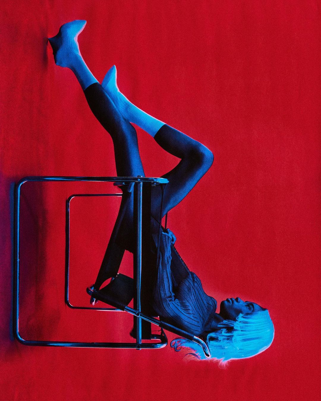 Upside-down fashion shot: Woman in blue tones poses with a metal chair against a vibrant red background in a studio setting.