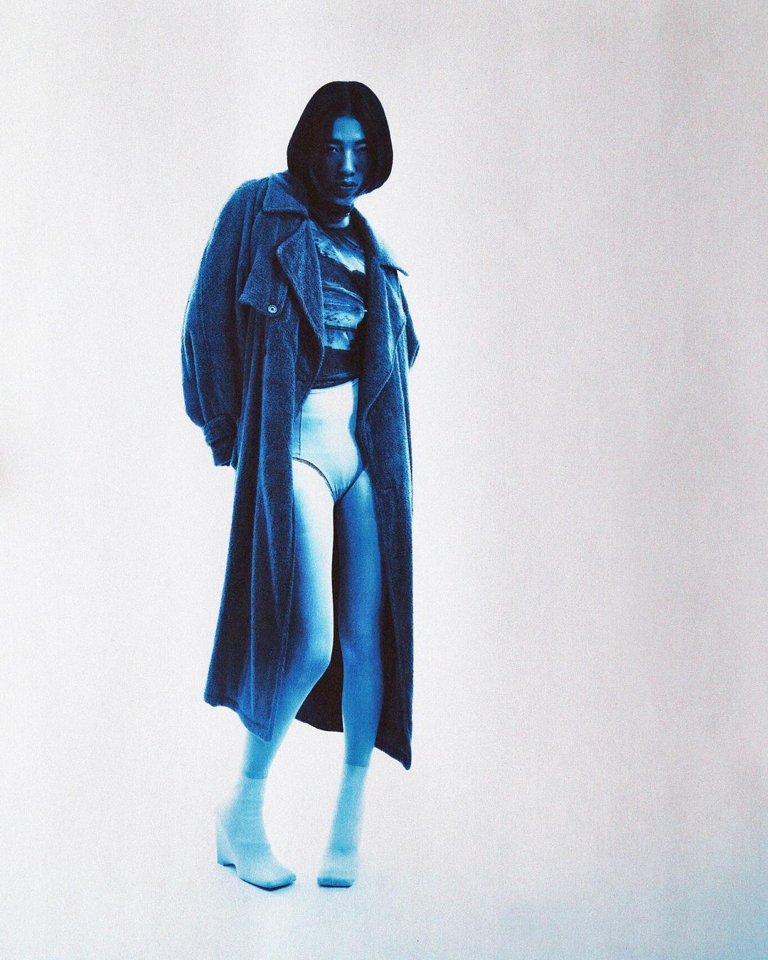 Fashionable woman in a blue-toned photo studio wearing a long coat, shorts, and boots against a clean white background.