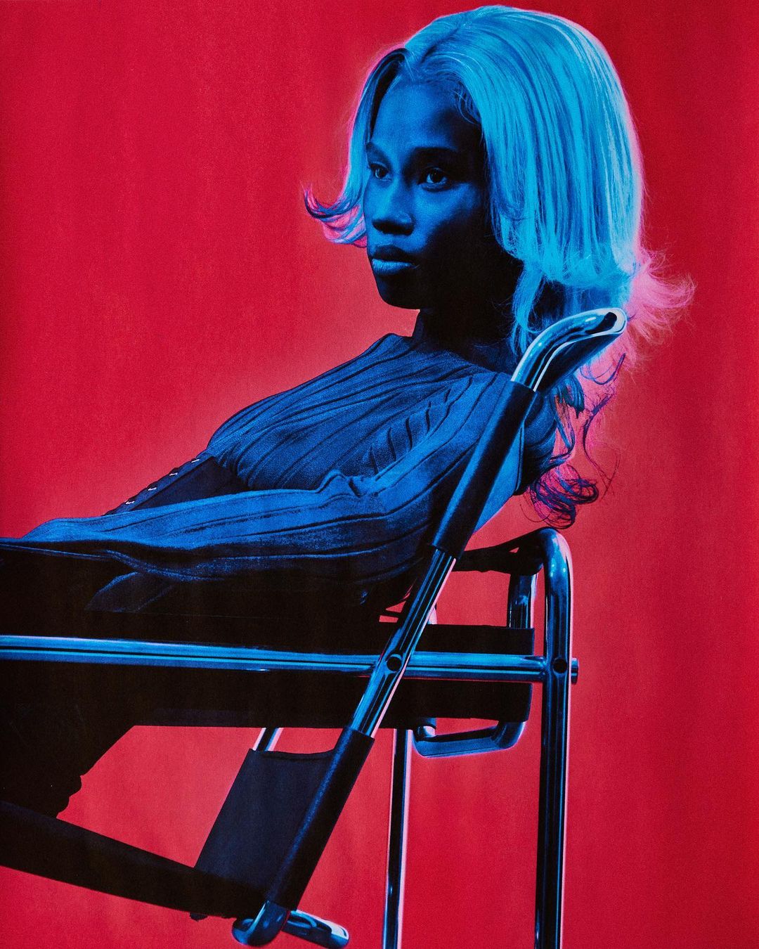 Blue-toned portrait of a Black woman with blonde hair sitting on a chair against a red backdrop in a studio environment.