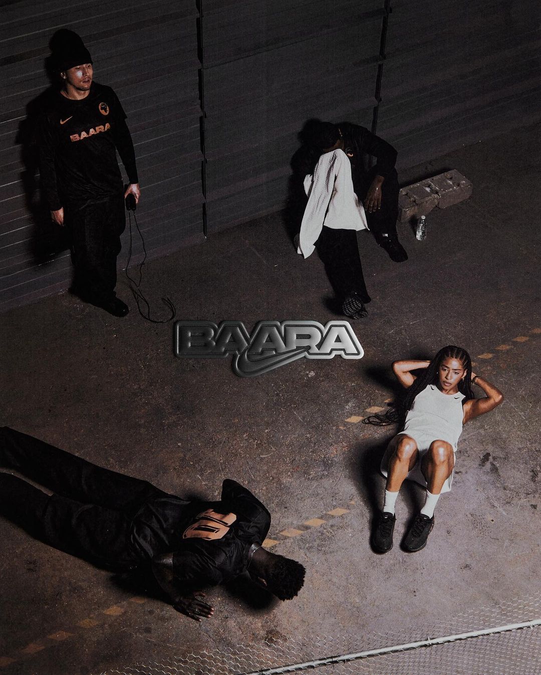 Four people pose in a warehouse-like indoor setting with a cement floor and gray walls under dim lighting, with BAARA text overlay.