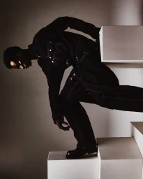 A Black man wearing a dark denim jumpsuit posing on white geometric blocks against a neutral studio backdrop.