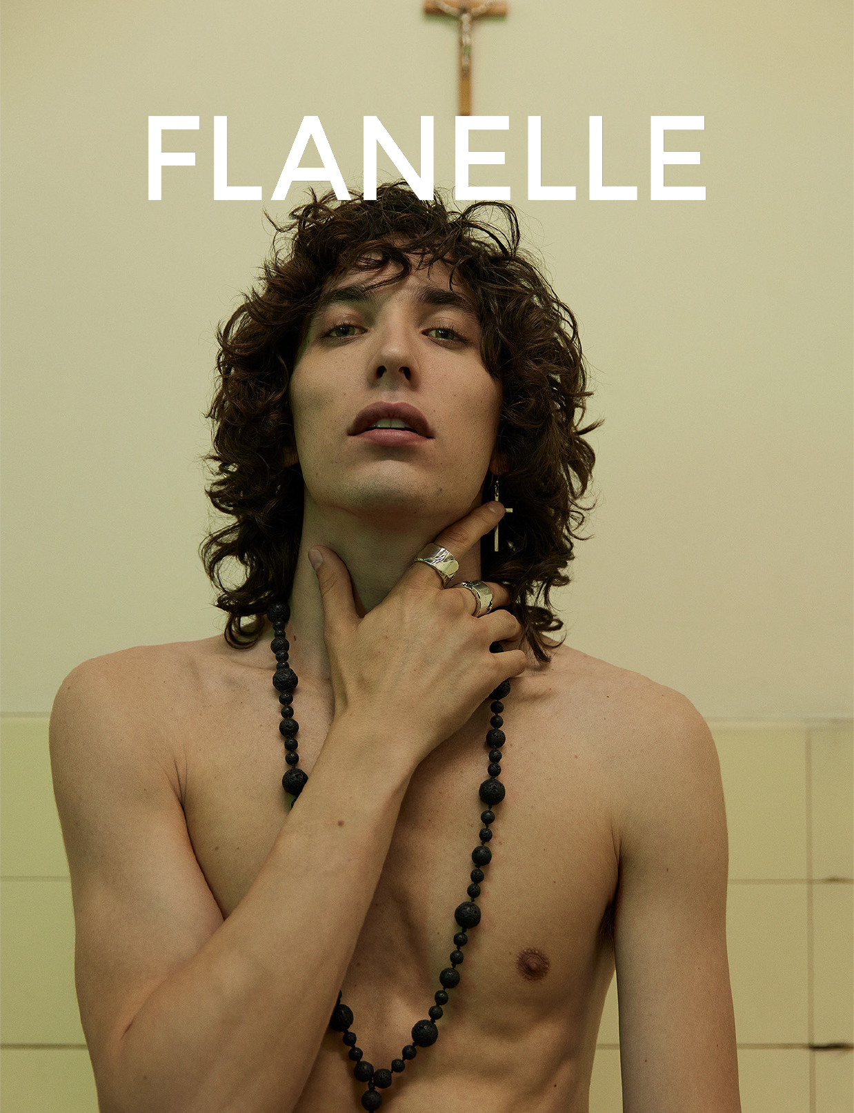 Portrait of a young adult male with curly hair, wearing a beaded necklace and cross earring, set against a tiled wall.