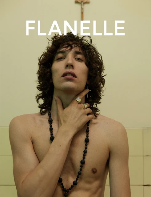 Portrait of a young adult male with curly hair, wearing a beaded necklace and cross earring, set against a tiled wall.
