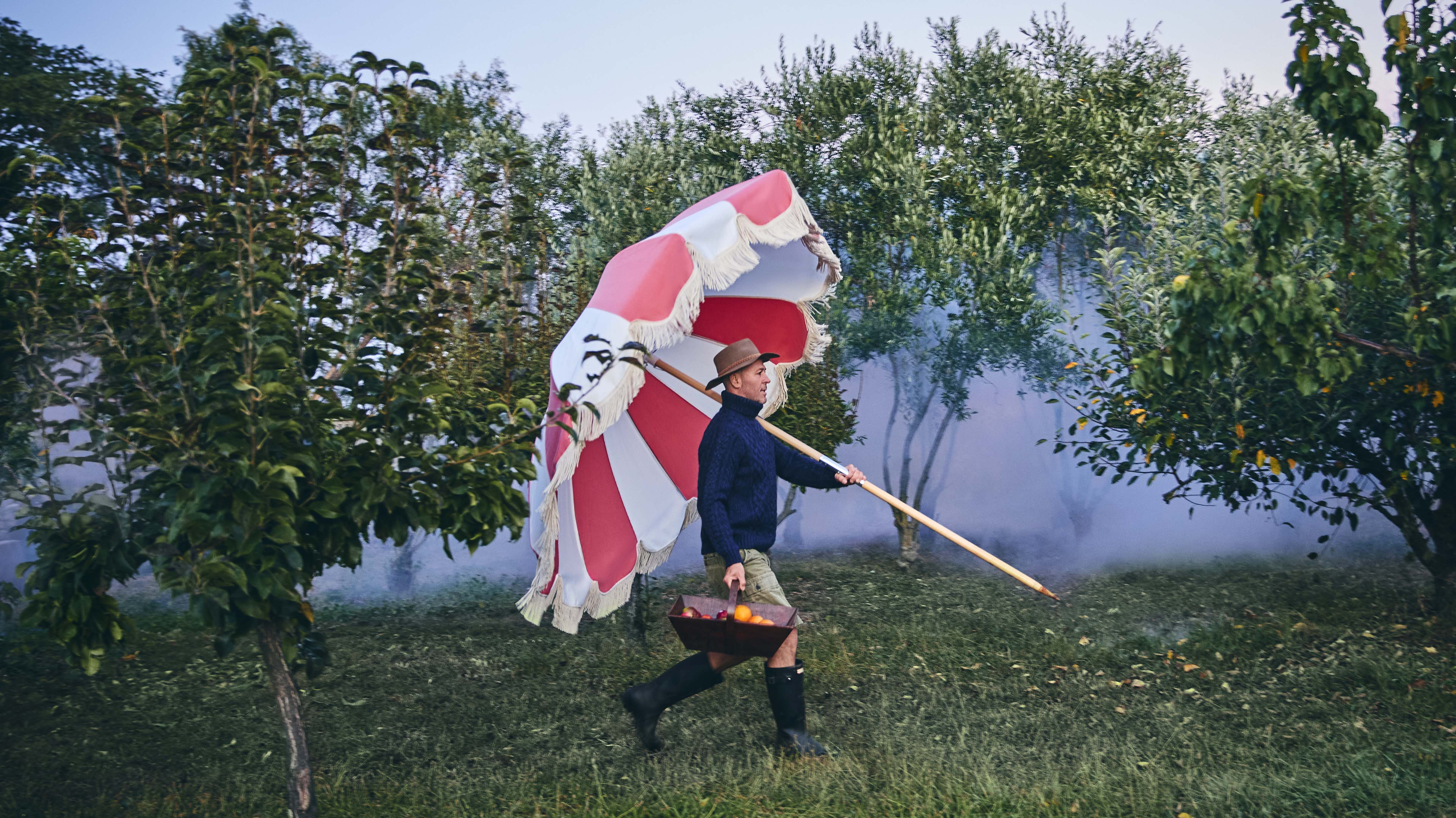 Man in hat carries red and white fringed parasol and fruit basket through green field with trees and light fog in the background.