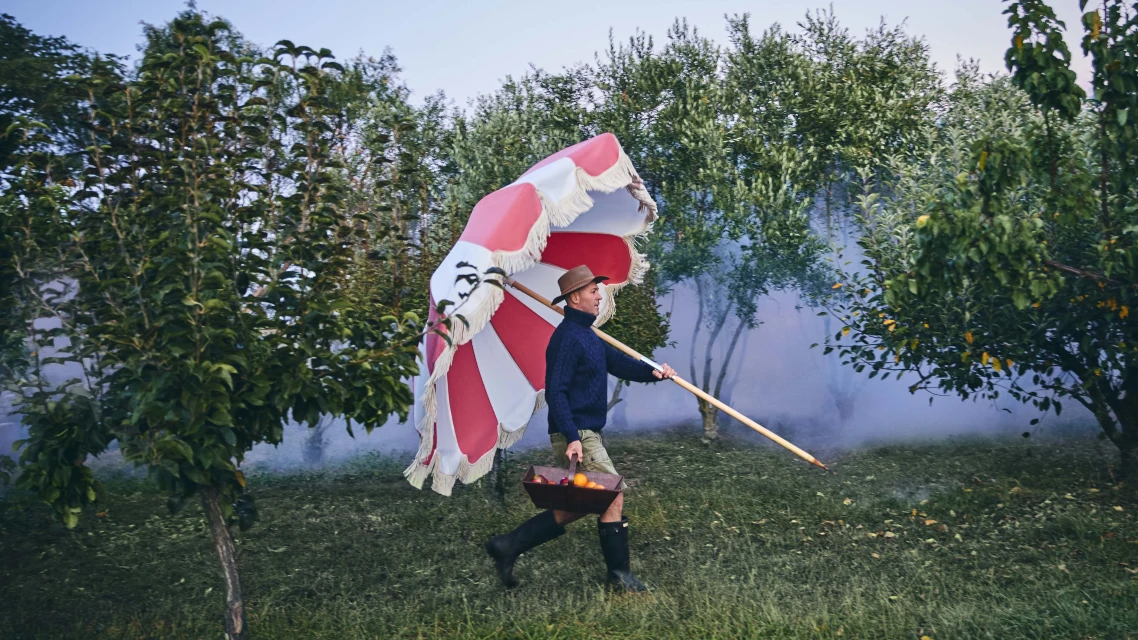 Man in hat carries red and white fringed parasol and fruit basket through green field with trees and light fog in the background.