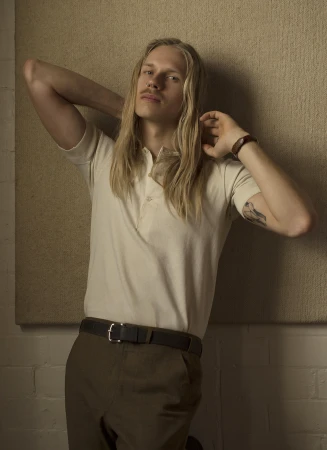 Portrait of a man with long blond hair wearing an off-white polo shirt and dark trousers against a textured panel background