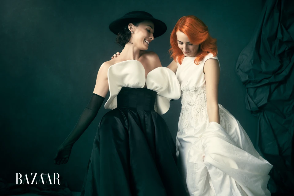 Two women in gowns pose in a fashion photo. One wears black and white with a hat, while the other wears white with red hair.