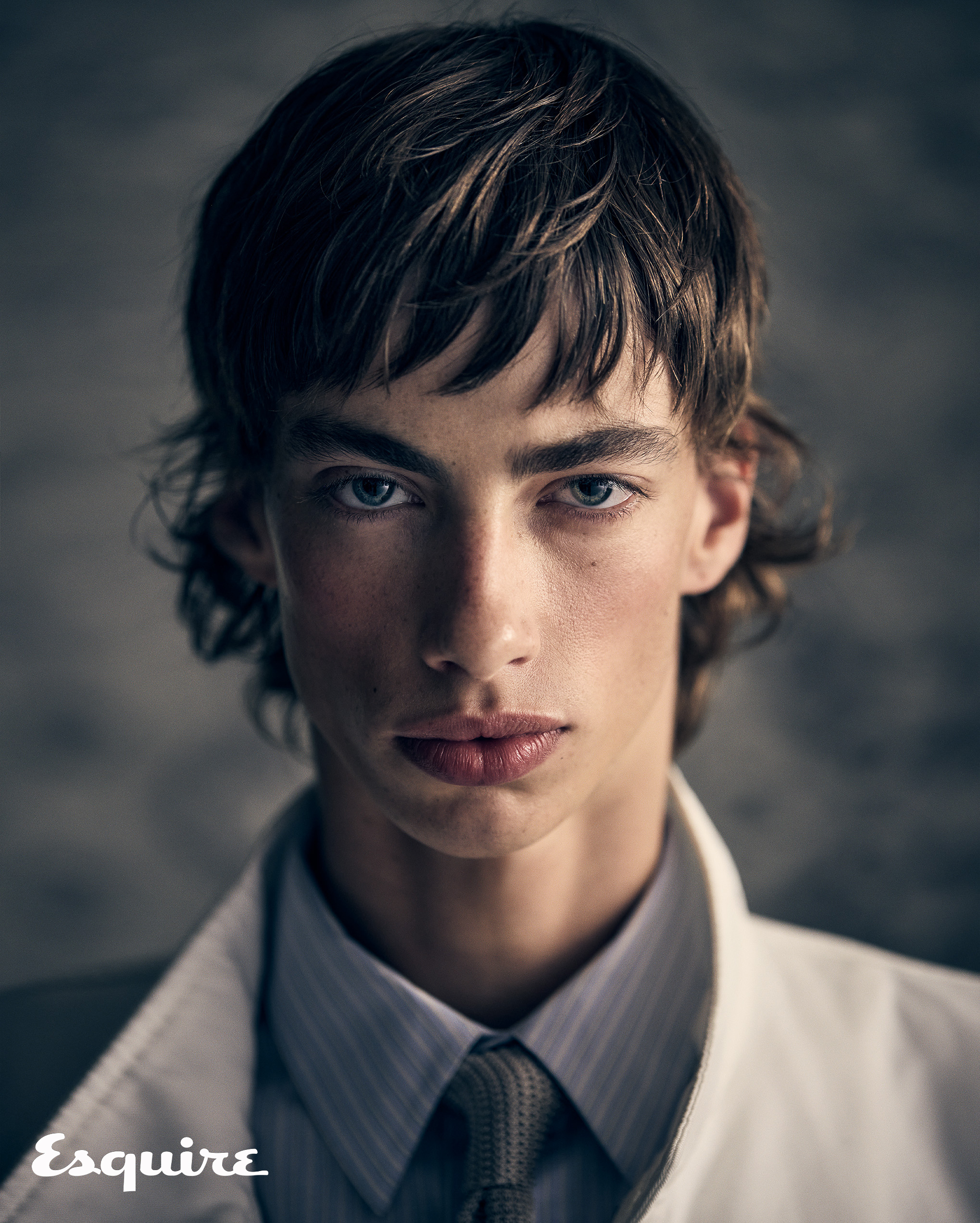 Close-up fashion portrait of a young adult male with brown hair wearing a white collared shirt and tie against a grey background.