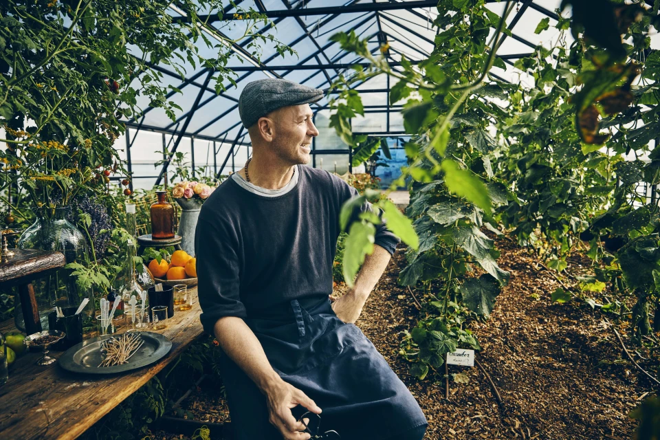 Man with cap sits in a greenhouse surrounded by plants. He is wearing an apron and looks to the side, with glasswares and fruits on a wood table.