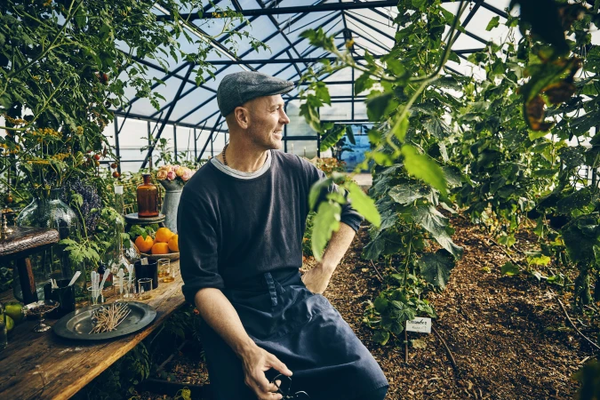 Man with cap sits in a greenhouse surrounded by plants. He is wearing an apron and looks to the side, with glasswares and fruits on a wood table.