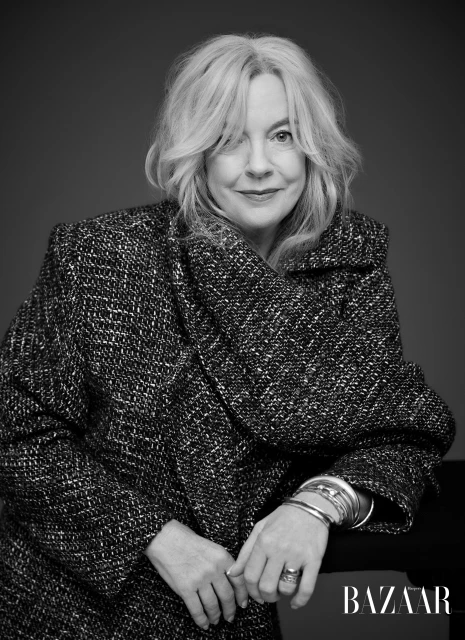 Black and white portrait of a mature woman with wavy blonde hair wearing a patterned jacket and silver bracelets against a dark background.
