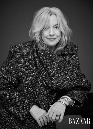Black and white portrait of a mature woman with wavy blonde hair wearing a patterned jacket and silver bracelets against a dark background.