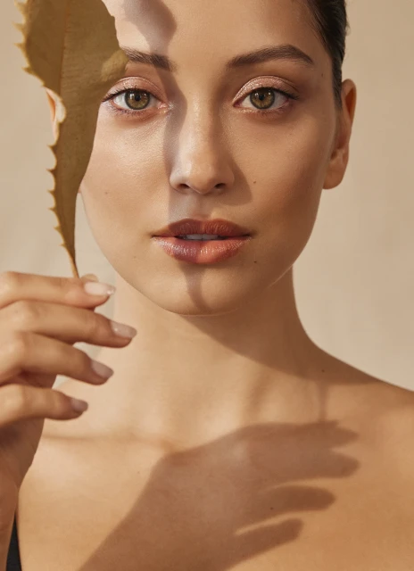 Close-up portrait of a young woman with fair skin, featuring a large leaf casting shadows across her face in a neutral-toned indoor setting.